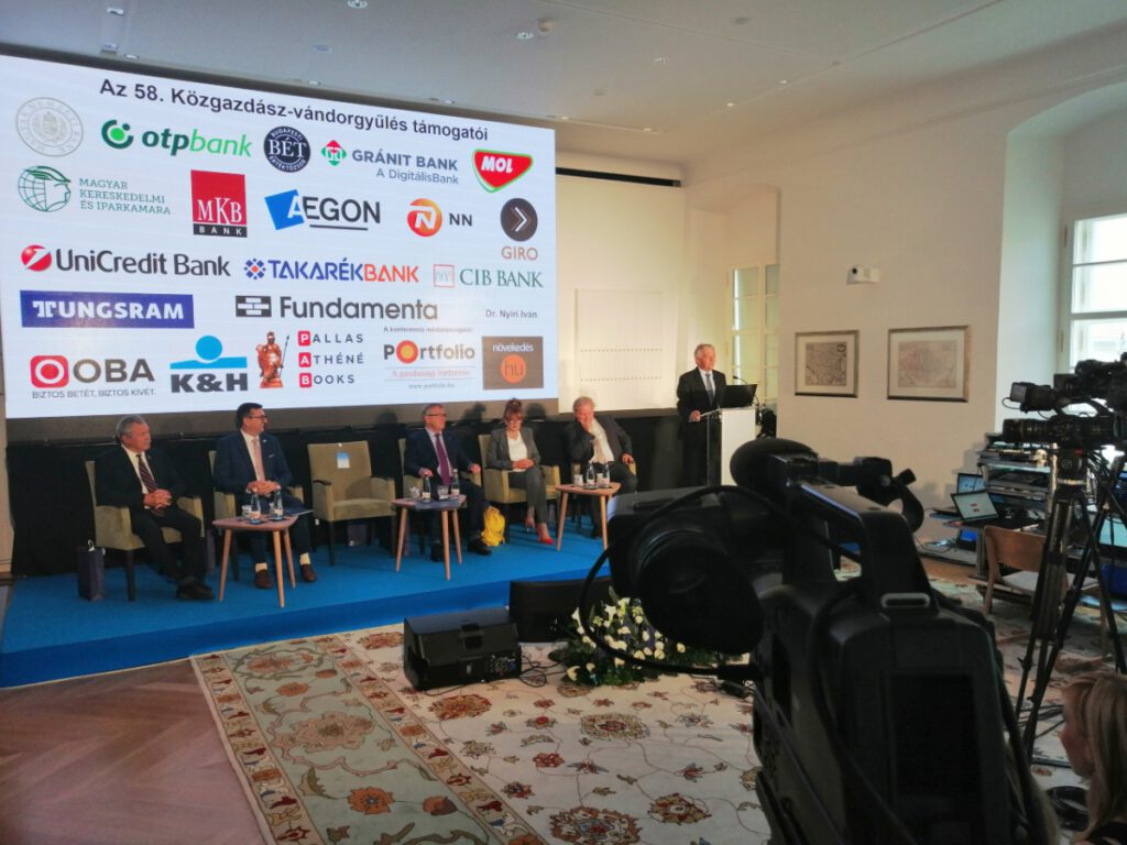 A képen az 58. Közgazdász Vándorgyűlés megnyitója látható, A színpadon az előadók a kivetítő előtt ülnek. Pleschinger Gyula pedig a mikrofon előtt áll és beszédet mond.
